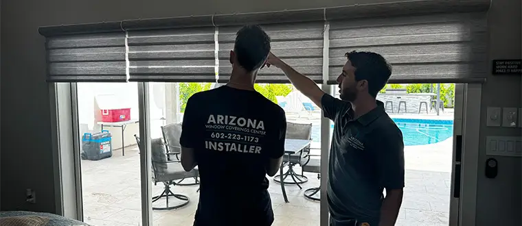 Two men stand indoors by large sliding glass doors with gray window shades. One points at the shades while the other, wearing an installer t-shirt with a company logo, listens intently. Through the Products Page view, a pool and patio furniture are visible outside. The man pointing wears a polo shirt.