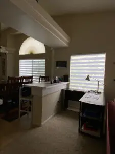 A dimly lit room features a bar-height counter with chairs and a dining table on the left. A desk lamp and papers sit on a small desk by the window with the best blinds installation near me. An arch-shaped window above allows soft light. A plush carpet covers the floor, creating a cozy atmosphere.