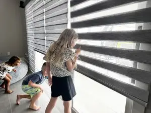 Three children stand by a large window with custom gray striped blinds. A girl with long hair peers through the blinds, while two younger children crouch and look outside. The room is softly lit, and the floor is tiled. The scene conveys curiosity and anticipation.