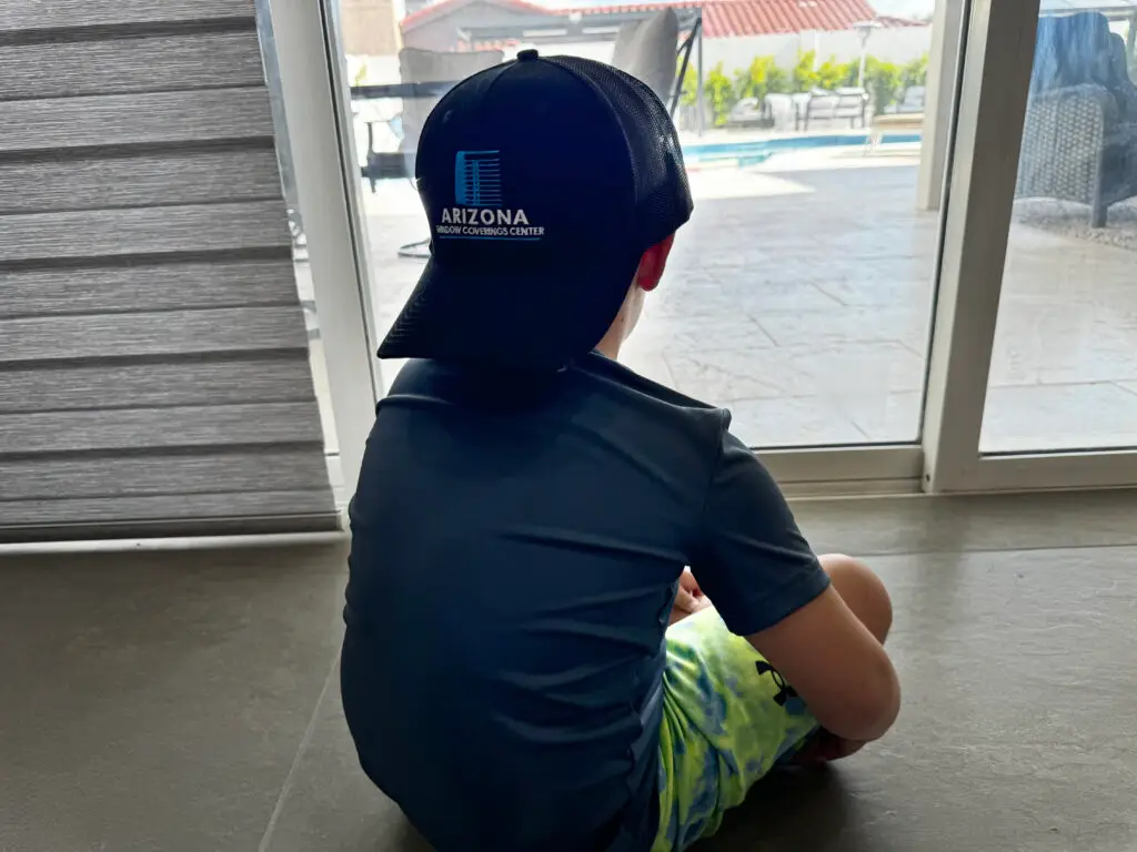 A child sits on the floor, facing a large window adorned with horizontal blinds. They are wearing a black cap with Arizona Window and Door Store written on it, a dark blue shirt, and tie-dye shorts. Outside the window, a pool and patio furniture are visible under a clear sky.